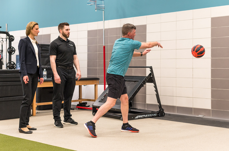 A man throwing an exercise ball at a wall while two people watch